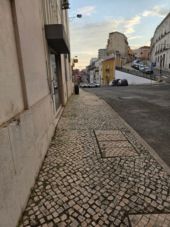 A Lisbon street with a sidewalk of small stone cobbles, parked cars and a road curving uphill between older buildings