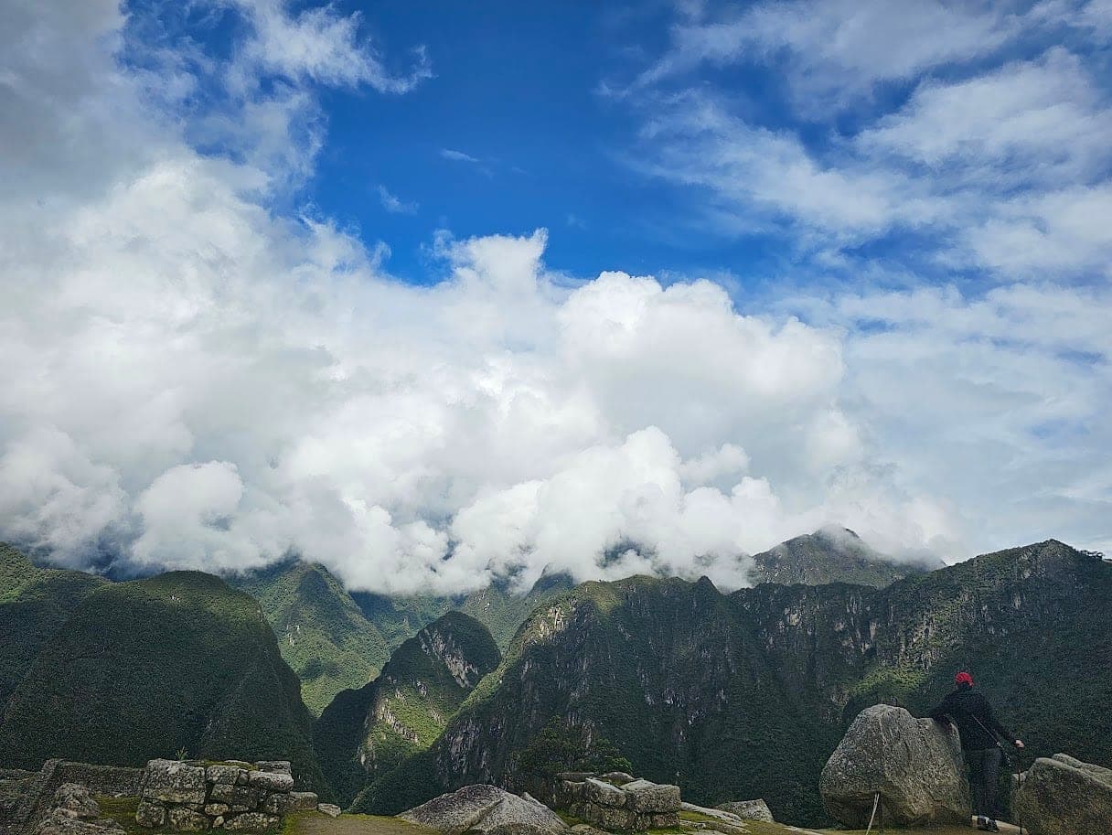 Cloud-wrapped green peaks seen from Machu Picchu, with Inca stone walls in the foreground and a hiker looking out on the right