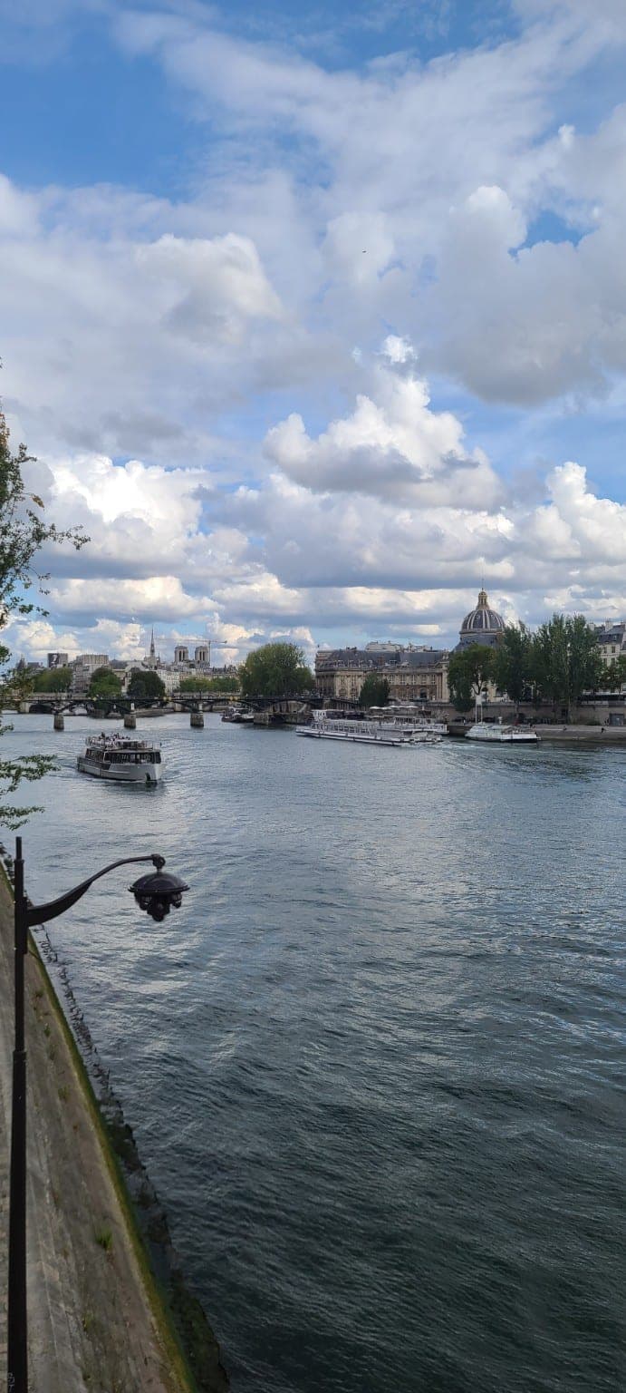 Boats on the Seine beneath a cloudy blue sky, with the Institut de France dome on the riverbank in Paris